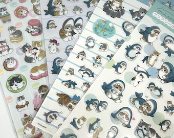 May include: Four sheets of clear stickers featuring mofusand cat illustrations. The designs include cats wearing shark costumes, cats interacting with sea creatures, and cats with various food items like macarons and cakes, all on transparent backing.
