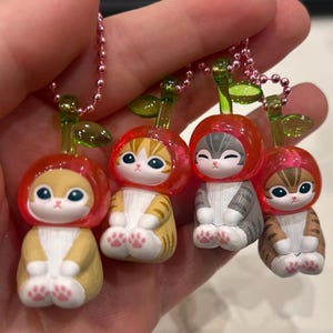 Pu&ograve; includere: Quattro piccole statuette di gatto, ognuna con un cappello rosso a forma di ciliegia e un gambo verde. I gatti hanno diversi motivi di pelliccia: giallo, arancione, grigio e marrone. Ogni statuetta &egrave; attaccata a una catena di perline rosa.