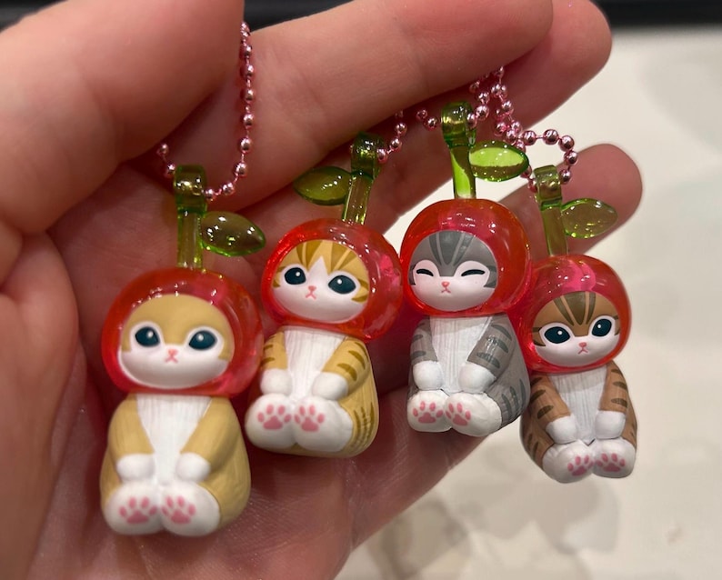 Pu&ograve; includere: Quattro piccole statuette di gatto, ognuna con un cappello rosso a forma di ciliegia e un gambo verde. I gatti hanno diversi motivi di pelliccia: giallo, arancione, grigio e marrone. Ogni statuetta &egrave; attaccata a una catena di perline rosa.