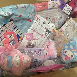 Peut inclure: Une boîte en carton remplie de divers produits dérivés des personnages Sanrio. Les articles comprennent des peluches, des pochettes, des porte-clés et des autocollants, avec des personnages comme Hello Kitty et My Melody. Les produits sont de couleurs rose, bleu et blanc.