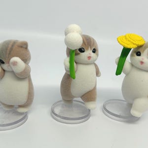 K&ouml;nnte beinhalten: Drei Katzenfiguren mit braun-wei&szlig;em Fell, die jeweils eine Blume halten. Eine Katze h&auml;lt ein gr&uuml;nes Kleeblatt, eine andere einen wei&szlig;en Wattebausch und die letzte eine gelbe Blume. Sie stehen auf transparenten, kreisf&ouml;rmigen Sockeln. H&ouml;he ca. 5 cm.
