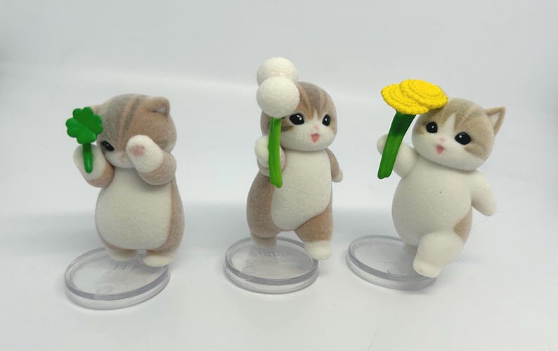 K&ouml;nnte beinhalten: Drei Katzenfiguren mit braun-wei&szlig;em Fell, die jeweils eine Blume halten. Eine Katze h&auml;lt ein gr&uuml;nes Kleeblatt, eine andere einen wei&szlig;en Wattebausch und die letzte eine gelbe Blume. Sie stehen auf transparenten, kreisf&ouml;rmigen Sockeln. H&ouml;he ca. 5 cm.