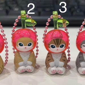 Pu&ograve; includere: Quattro statuette di gatto con cappelli rosa a forma di ciliegia e portachiavi. I gatti hanno diversi motivi di pelliccia: tigrato, marrone chiaro e grigio. Ogni statuetta ha una catena di perline rosa e un gambo verde.