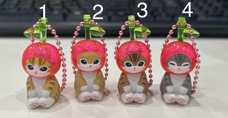 Pu&ograve; includere: Quattro statuette di gatto con cappelli rosa a forma di ciliegia e portachiavi. I gatti hanno diversi motivi di pelliccia: tigrato, marrone chiaro e grigio. Ogni statuetta ha una catena di perline rosa e un gambo verde.