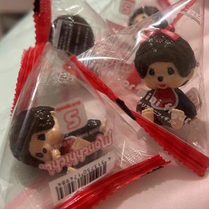 Peut inclure: Deux figurines miniatures Monchhichi emballées dans des sachets triangulaires en plastique transparent avec des bords rouges scellés. Chaque figurine a des cheveux brun foncé, un nœud rouge, un petit nez et un bavoir rouge.