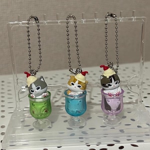 Puede incluir: Tres llaveros con temática de gatos. Cada llavero presenta una pequeña figura de gato dentro de un vaso colorido. Los gatos llevan pequeños sombreros con una cereza encima. Los vasos son verdes, azules y morados, y están unidos a cadenas de bolas plateadas.