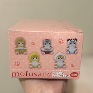 May include: A pink box featuring illustrations of cat figurines in various costumes. The box has the brand name "mofusand" and Japanese text. The figurines are in different outfits, including a panda, a hedgehog, and a hooded cat.