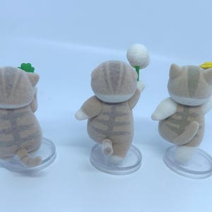 K&ouml;nnte beinhalten: Drei kleine, hellbraune Katzenfiguren mit gestreiften Mustern und wei&szlig;en Akzenten. Jede Katze h&auml;lt einen anderen Gegenstand: ein gr&uuml;nes Kleeblatt, einen wei&szlig;en Ball und eine gelbe Blume. Sie stehen auf transparenten, kreisf&ouml;rmigen Sockeln.
