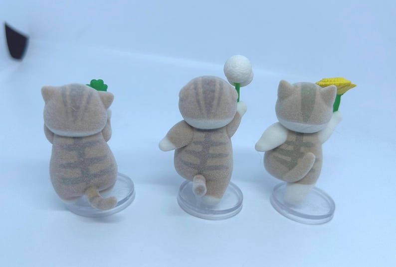 K&ouml;nnte beinhalten: Drei kleine, hellbraune Katzenfiguren mit gestreiften Mustern und wei&szlig;en Akzenten. Jede Katze h&auml;lt einen anderen Gegenstand: ein gr&uuml;nes Kleeblatt, einen wei&szlig;en Ball und eine gelbe Blume. Sie stehen auf transparenten, kreisf&ouml;rmigen Sockeln.