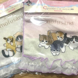 May include: Two packaged hand towels featuring the "mofusand" brand. One towel has a light background with cat illustrations in yellow and black outfits. The other towel has a pink background with multiple cat illustrations. Both towels have a ruffled trim.