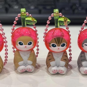 Pu&ograve; includere: Quattro statuette di gatto con cappelli a forma di ciliegia. I gatti hanno diversi motivi di pelliccia: marrone chiaro, marrone e grigio. Ogni statuetta ha un cappello a ciliegia rosa, una catena di perline rosa e un gambo verde. Le statuette sono su una superficie bianca.