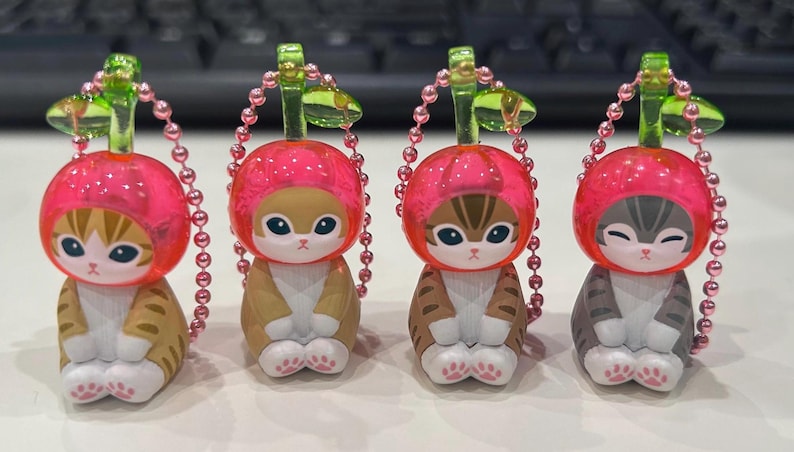 Pu&ograve; includere: Quattro statuette di gatto con cappelli a forma di ciliegia. I gatti hanno diversi motivi di pelliccia: marrone chiaro, marrone e grigio. Ogni statuetta ha un cappello a ciliegia rosa, una catena di perline rosa e un gambo verde. Le statuette sono su una superficie bianca.