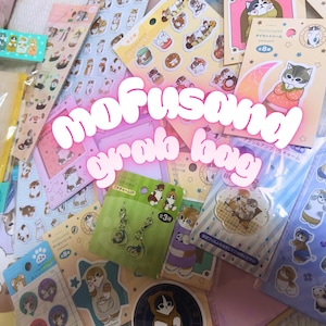 Op de afbeelding: Een verzameling stickers, bedels en andere items met cartoonkatten en -honden. De afbeelding bevat de tekst "mofusand grab bag" in een roze, bubbelachtige lettertype, en diverse sticker vellen en bedels.