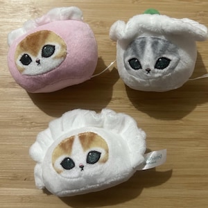 May include: Three plush cat-faced dumplings in pink, white, and white. Each dumpling features an embroidered cat face with large eyes and a small nose. The white dumplings have a tag that reads "fusand".