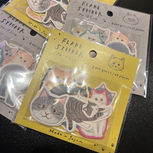 May include: Multiple packs of cat-themed flake stickers. Each pack features diverse cat designs in various colours and patterns. The packaging includes Japanese text and the phrase "Made in Japan."
