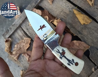May include: A handcrafted knife with a polished blade featuring a brown animal silhouette and the initials "CG". The handle is off-white with a black and red decorative design. The "HandArts Cutlery" logo is visible in the upper left corner.