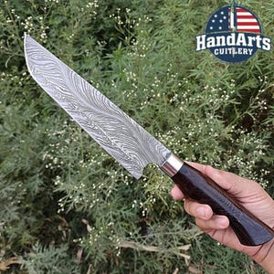 Puede incluir: Un cuchillo de chef de acero de Damasco con mango de madera oscura. La hoja presenta un patrón de ondas único. El cuchillo se muestra sobre un fondo de follaje verde. El logotipo "HandArts Cutlery" es visible en la esquina superior derecha.