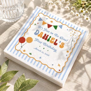 May include: A stack of white paper napkins with a blue striped border and a decorative orange and yellow scalloped edge. The napkins feature the text "It's Fun to Be One! DANIEL'S First Birthday March 12, 2026" with colorful balloons and stars.