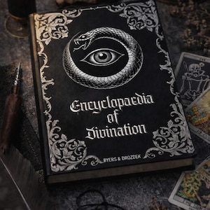Op de afbeelding: Een zwart boek getiteld "Encyclopaedia of Divination" met een slang- en oogontwerp. Het boek is omgeven door tarotkaarten, een brandende kaars en een pentagramhanger. De kaft van het boek heeft sierlijke zilveren details.