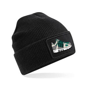 May include: A black ribbed knit beanie featuring a rectangular patch on the front. The patch displays a graphic illustration of a sneaker in shades of grey, white, and green with black stripes on the side, set against a solid black background.