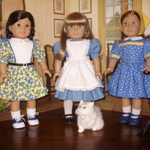 May include: Three dolls wearing different dresses and shoes. The doll on the left is wearing a yellow and blue floral dress with a white collar and navy blue shoes. The doll in the middle is wearing a blue and white dress with a white apron and white lace trim. The doll on the right is wearing a blue and yellow polka dot dress with a yellow collar and navy blue shoes.