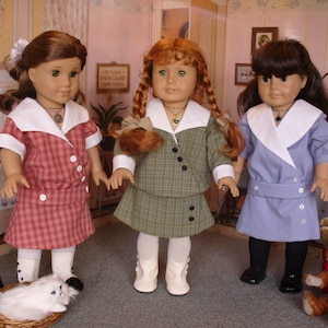 May include: Three dolls wearing different plaid dresses with white collars. The dolls have brown, red, and blonde hair. The dolls are standing in a room with a pink wall and a white window.