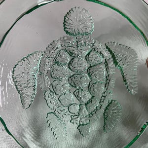 Puede incluir: Un cuenco redondo de vidrio transparente con un diseño de tortuga marina en relieve en el centro. La tortuga tiene aletas y caparazón texturizados. El cuenco tiene un borde festoneado y un ligero tinte verde. Diámetro aproximado de 25 cm.