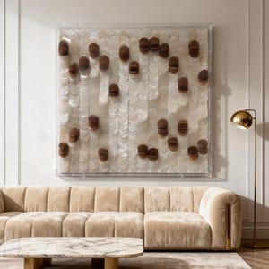 Op de afbeelding: Een groot, vierkant kunstwerk voor aan de muur met een helder acryl frame. Het kunstwerk heeft een patroon van witte, schelpachtige elementen afgewisseld met bruine, cilindrische accenten. Een beige hoekbank en marmeren salontafel staan op de voorgrond.