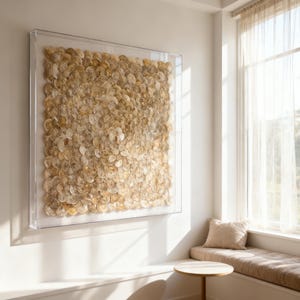 Op de afbeelding: Een ingelijst kunstwerk met een mozaïek van kleine, ronde, beige schelpen. De schelpen zijn in een dicht patroon gerangschikt in een heldere acryl lijst. Het kunstwerk hangt aan een witte muur, dichtbij een raam met transparante gordijnen.