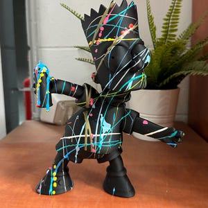 Peut inclure: Figurine noire d'un personnage de dessin animé avec des éclaboussures de peinture colorées. La figurine tient une bombe de peinture et a une coiffure en forme de couronne. La figurine est posée sur une surface en bois.