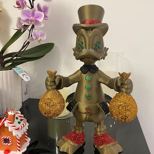 May include: A gold-colored figurine of a duck wearing a top hat with a red glitter band, holding two gold glitter money bags. The duck has green dollar sign eyes and a black bow tie. The figurine's feet are red glitter.