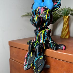 May include: A black Minnie Mouse figurine with a blue bow and colorful paint splatter design. The figurine is sitting on a wooden dresser. The paint colors include pink, yellow, and green.