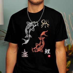 以下が含まれることがあります： 黒いTシャツ。白とオレンジの鯉のデザインと、日本のスタイルのイラストが特徴です。Tシャツには日本語の文字と山のグラフィックが含まれています。デザインは黒いTシャツにプリントされています。