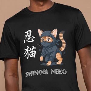以下が含まれることがあります： 黒いTシャツ。青い忍者衣装とマスクを着た漫画の忍者猫のデザイン。シャツには日本語の文字と白い「SHINOBI NEKO」の文字が入っています。