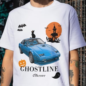 以下が含まれることがあります： 青いスポーツカー、黒猫、コウモリ、カボチャ、お化け屋敷、幽霊が描かれた白いTシャツ。「GHOSTLINE Classics」の文字が車の真下に黒でプリントされています。