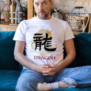 以下が含まれることがあります： 金と黒のドラゴンのデザインと黒で「DRAGON」という文字が入った白いTシャツ。青いジーンズと白い靴下を履いています。ターコイズ色のソファに座っています。