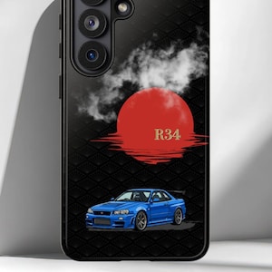 以下が含まれることがあります： 黒いスマートフォンケースで、下部に青いR34スポーツカーのグラフィックが描かれています。車の上には白い雲を伴う大きな赤い太陽と、金色のR34という文字があります。背景全体には、控えめな黒い扇形のパターンが施されています。