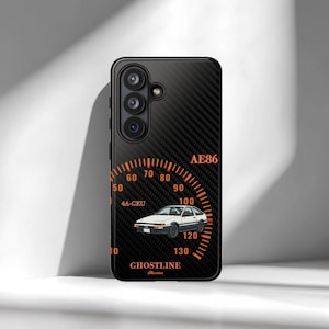 Könnte beinhalten: Schwarze Handyhülle mit Karbonfasertextur und orangefarbenem Tachometer-Design. Das Design beinhaltet eine weiße Autoillustration, den Text "AE86" und das Wort "GHOSTLINE". Die Hülle hat ein Kameramodul mit drei Objektiven.