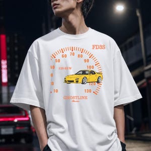 May include: A white short-sleeve t-shirt featuring a graphic print of a yellow sports car centered within an orange speedometer gauge design. The text FD3S, 13B-REW, and GHOSTLINE are printed in orange around the car illustration on the front.