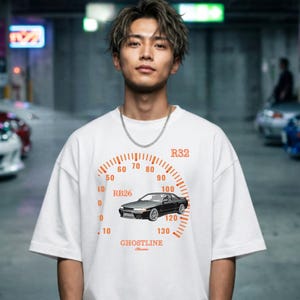 May include: A white short-sleeve t-shirt featuring a graphic print of a black R32 sports car centered within an orange speedometer gauge design. The graphic includes the text R32, RB26, and GHOSTLINE printed in orange on the front of the shirt.