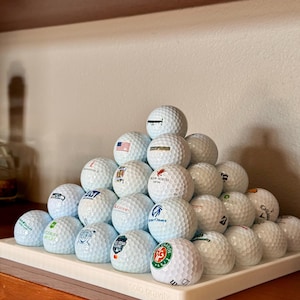 Op de afbeelding: Een piramide van witte golfballen, elk met een ander logo, rust op een wit rechthoekig dienblad. Het dienblad staat op een houten oppervlak. De tekst "solo gravity" is op het dienblad gedrukt.