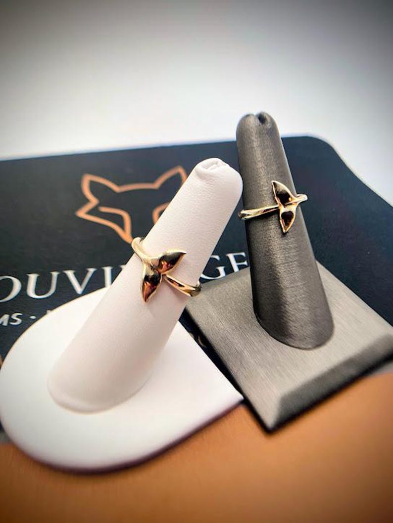 May include: Two gold rings, each featuring a delicate whale tail design. The rings are displayed on individual finger-shaped stands, one white and one dark gray, showcasing the polished metallic finish and the curved shape of the whale tail motif.