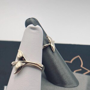 May include: Two silver-toned rings, each featuring a whale tail design. The rings are displayed on two separate finger-shaped stands, one white and one dark gray, showcasing the metallic finish and the curved shape of the whale tail motif.