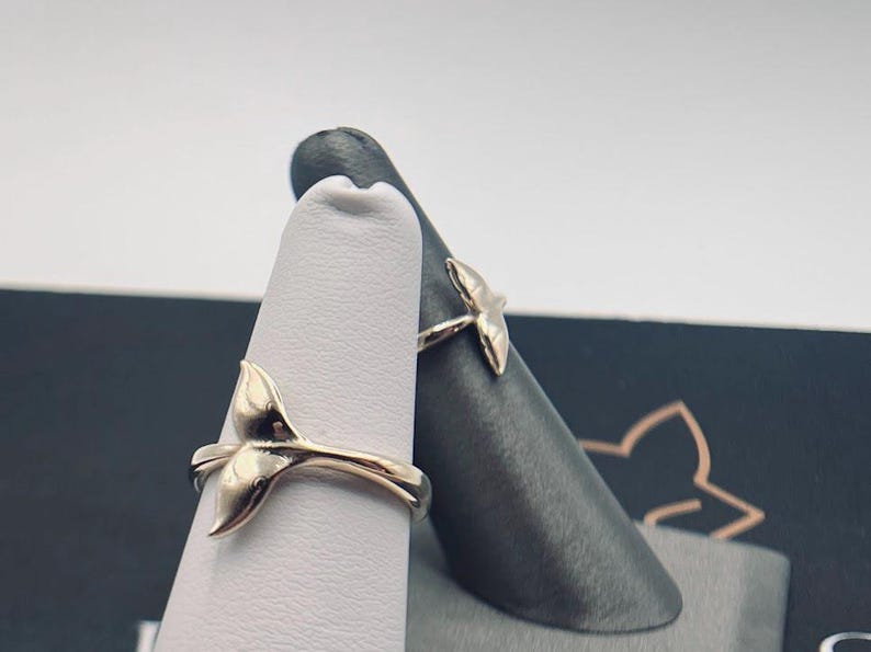 May include: Two silver-toned rings, each featuring a whale tail design. The rings are displayed on two separate finger-shaped stands, one white and one dark gray, showcasing the metallic finish and the curved shape of the whale tail motif.