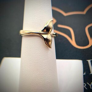 May include: A polished gold-tone ring featuring a prominent whale tail design as the centerpiece. The band is smooth and slender, connecting to the base of the tail. The metal surface reflects light, highlighting the curved, organic shape of the fluke.