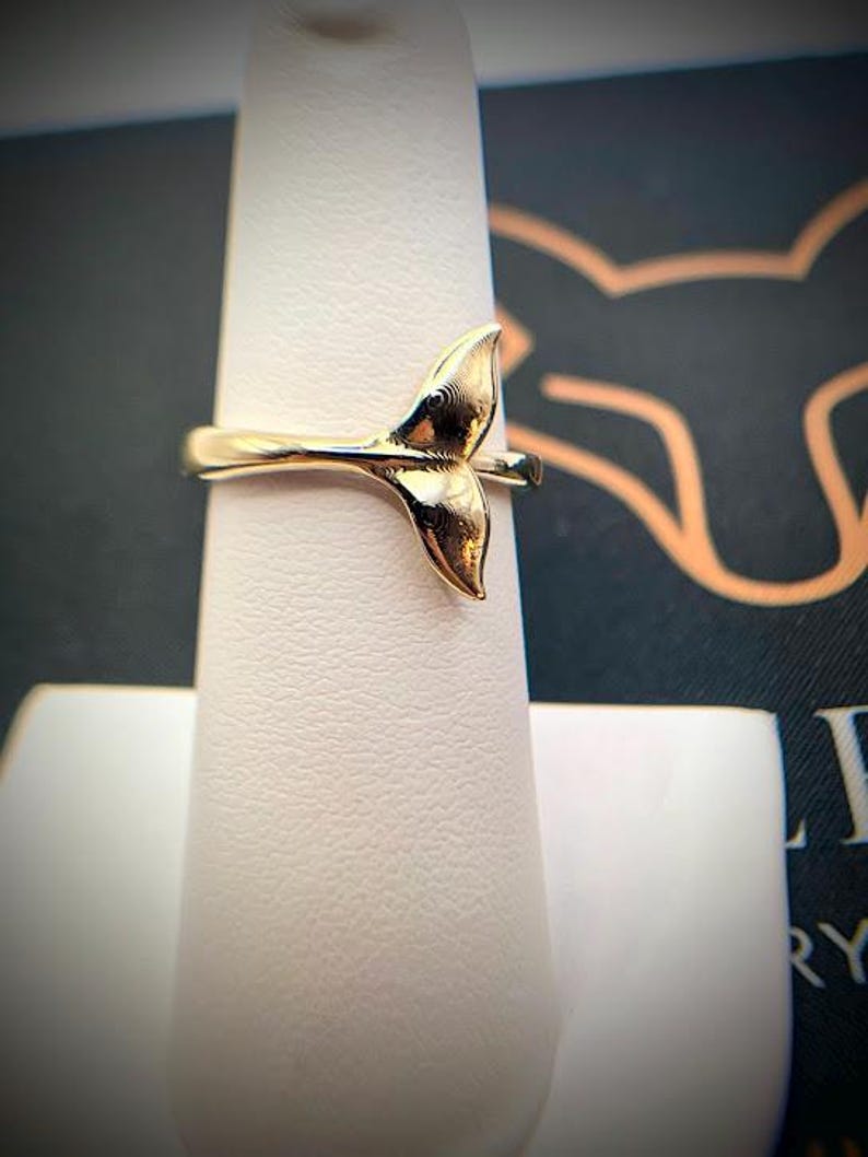 May include: A polished gold-tone ring featuring a prominent whale tail design as the centerpiece. The band is smooth and slender, connecting to the base of the tail. The metal surface reflects light, highlighting the curved, organic shape of the fluke.