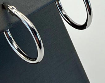 May include: A pair of polished silver-toned hoop earrings featuring a smooth, rounded metal construction. Each earring is designed with a hinged post mechanism at the top, allowing the hoop to open and close securely for comfortable wear.