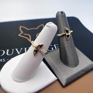 May include: Two gold-toned rings, each featuring a delicate whale tail design. The rings are displayed on individual finger-shaped stands, one white and one dark gray, showcasing the metallic finish and the curved shape of the tail motif.