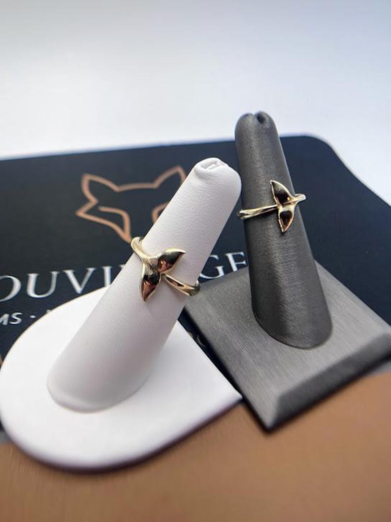 May include: Two gold-toned rings, each featuring a delicate whale tail design. The rings are displayed on individual finger-shaped stands, one white and one dark gray, showcasing the metallic finish and the curved shape of the tail motif.