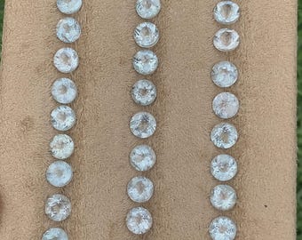 May include: A collection of forty-two small, round, faceted clear gemstones arranged in three vertical rows on a tan, textured display board. Each gemstone features a brilliant cut with multiple facets that reflect light, creating a sparkling appearance.